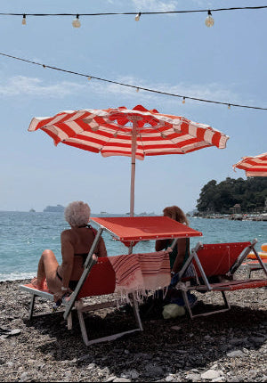 Two people sitting under red and white striped umbrellas on a beach.